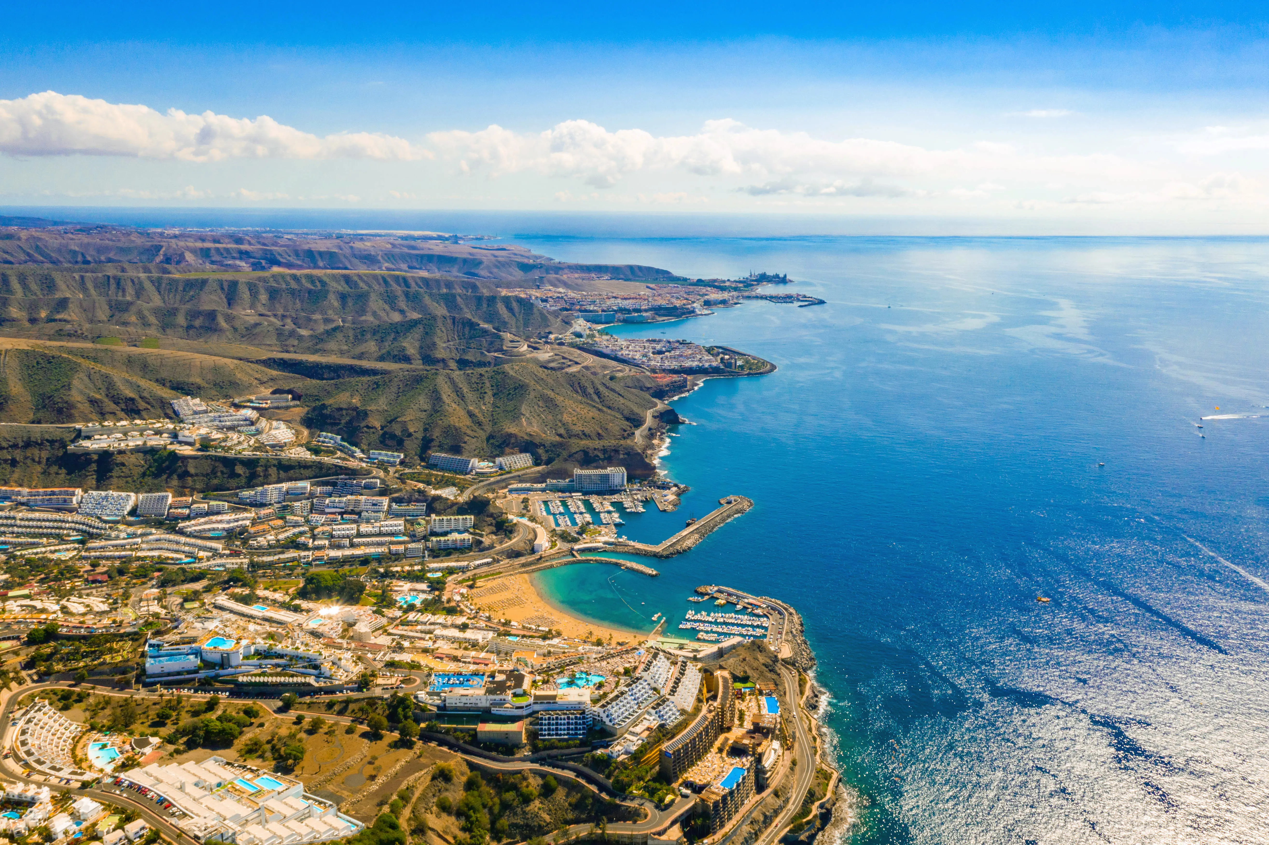 Luftfoto av Gran Canaria-kysten med havet og fjellene i bakgrunnen, viser turistområder og båthavn under en klar blå himmel