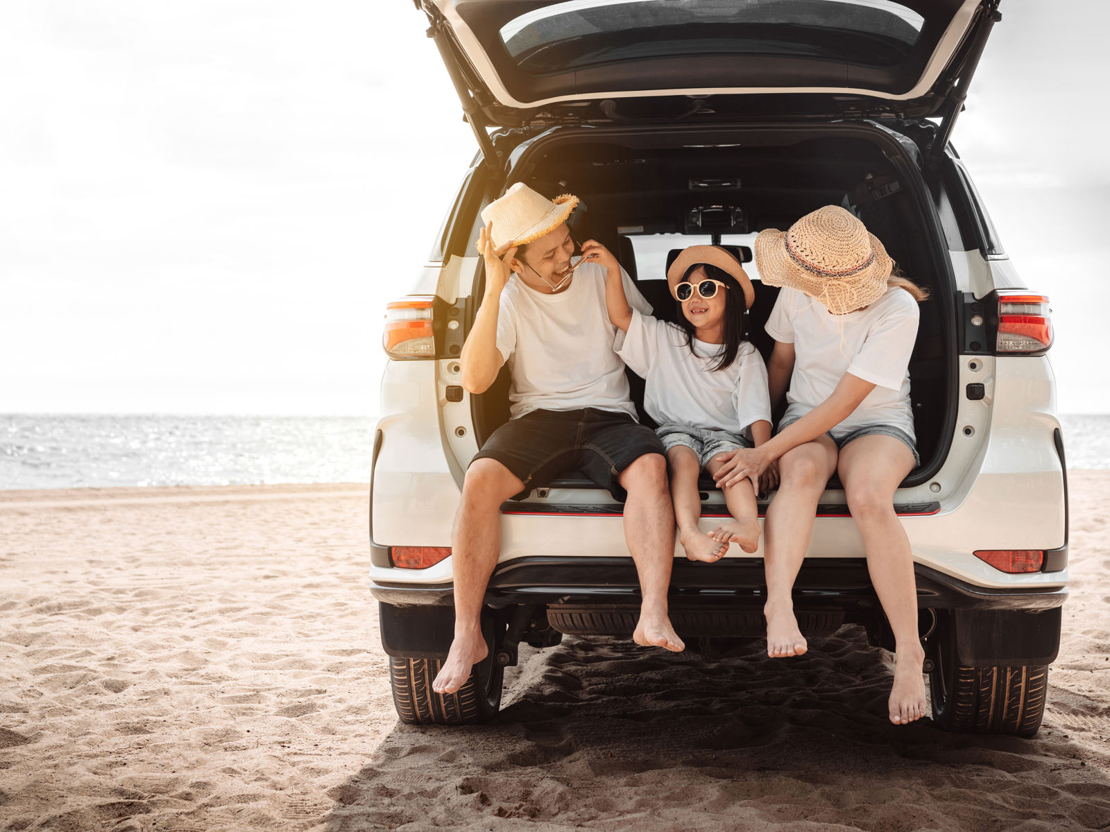 Compare travel insurance - Three people sitting in the trunk of an SUV by the beach, wearing sun hats and enjoying the holiday.