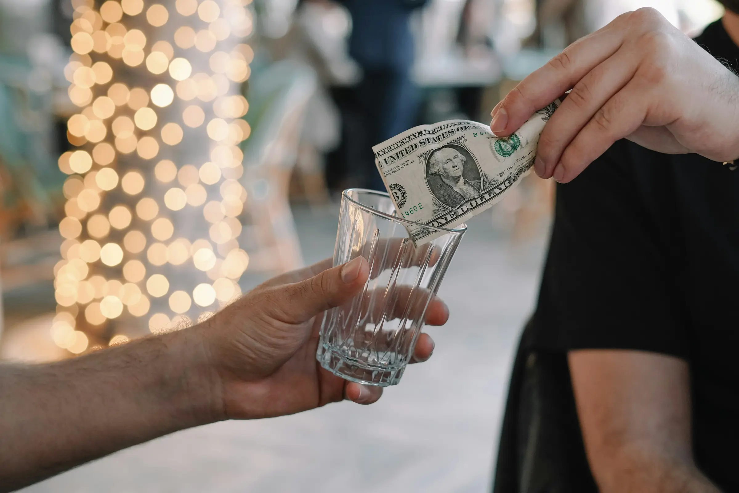 To personer deltar i en transaksjon med en én-dollarseddel som plasseres i et glass. Bakgrunnen er uskarp med dekorative, lyse bokeh-lys.