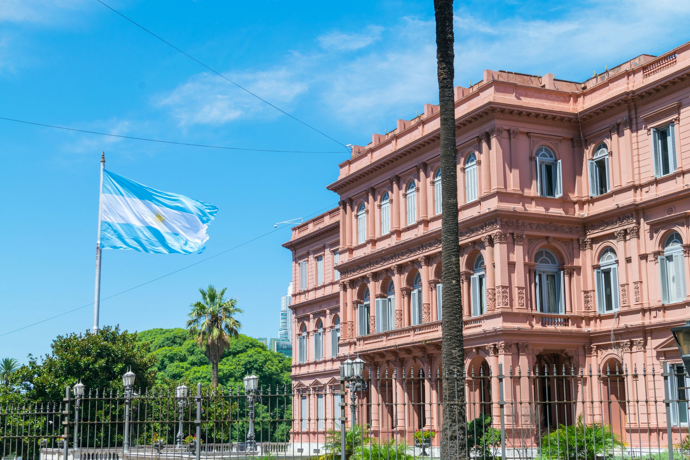 Argentinas flagg vaier foran presidentpalasset Casa Rosada i Buenos Aires under en klar blå himmel med grønt i forgrunnen.