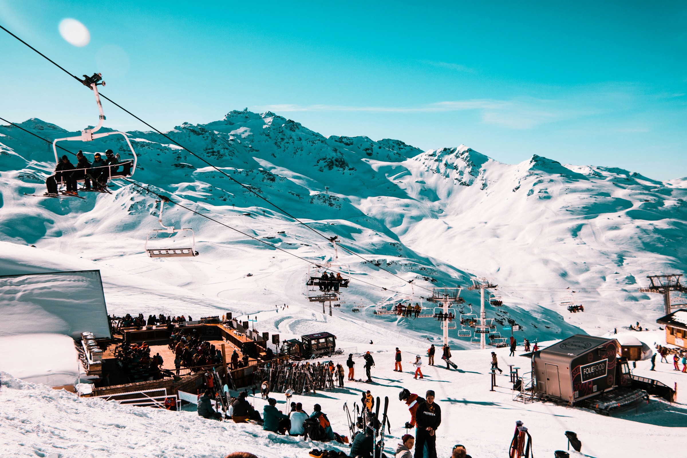 Lively ski resort with people on the ski lift and slopes, surrounded by snow-capped mountain peaks under a clear blue sky.