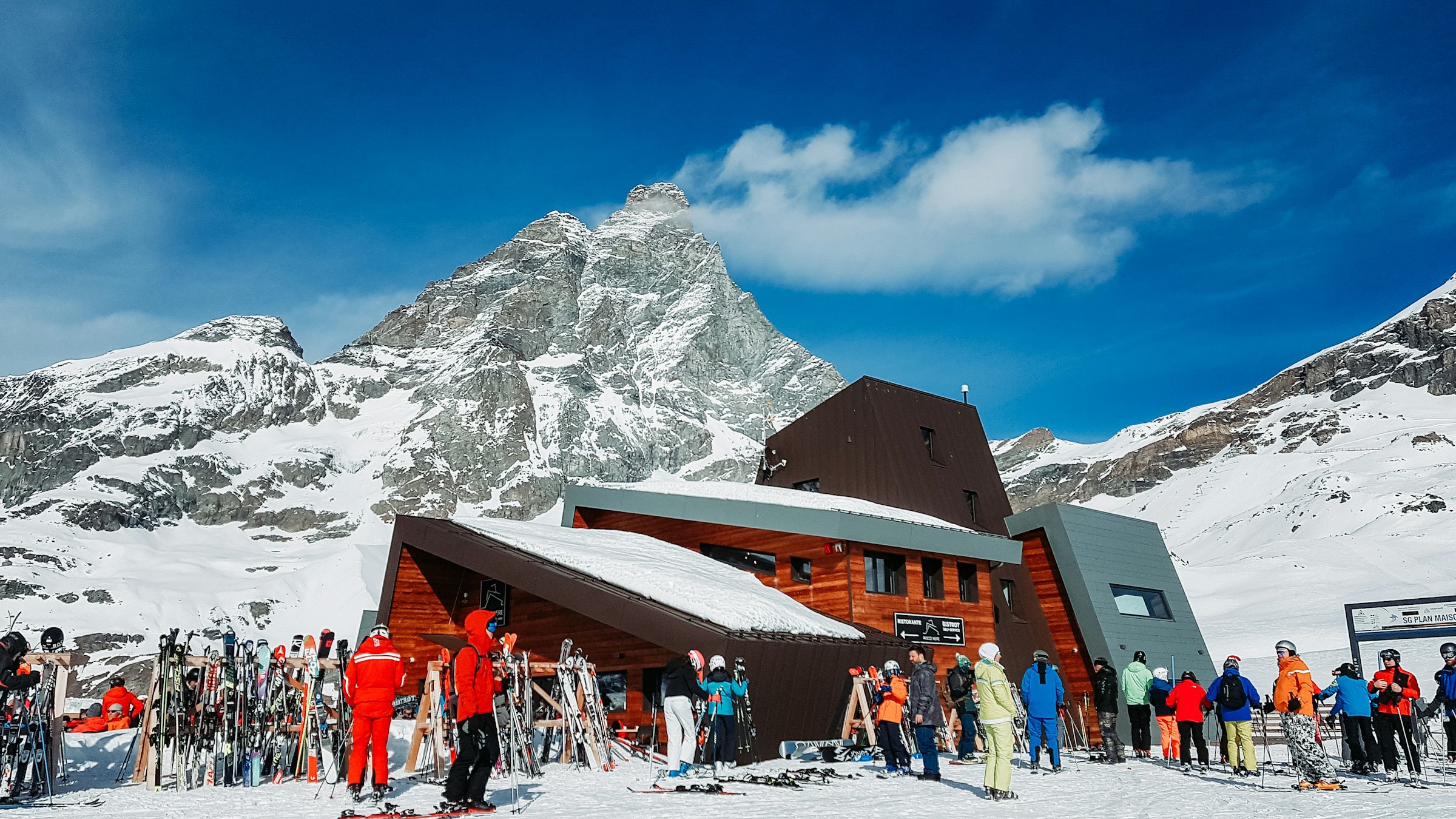 Skiers gather at a mountain hut in the alpine area with snow-capped mountain peaks in the background.