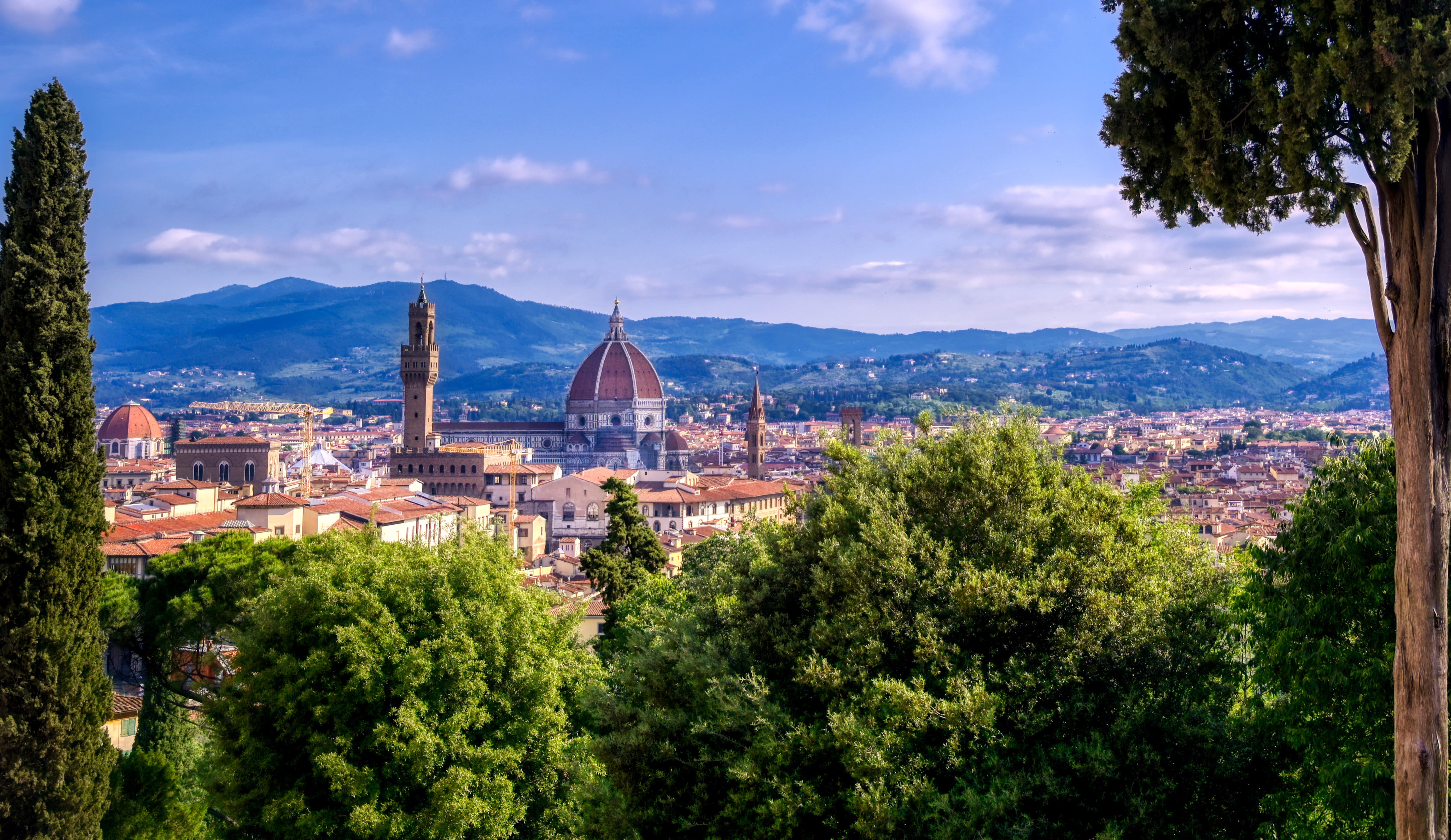 Reis til Firenze - Utsikt fra en høyde over Firenze i Italia hvor frodige trær møter byen med berømt kirke og blå himmel i bakgrunnen