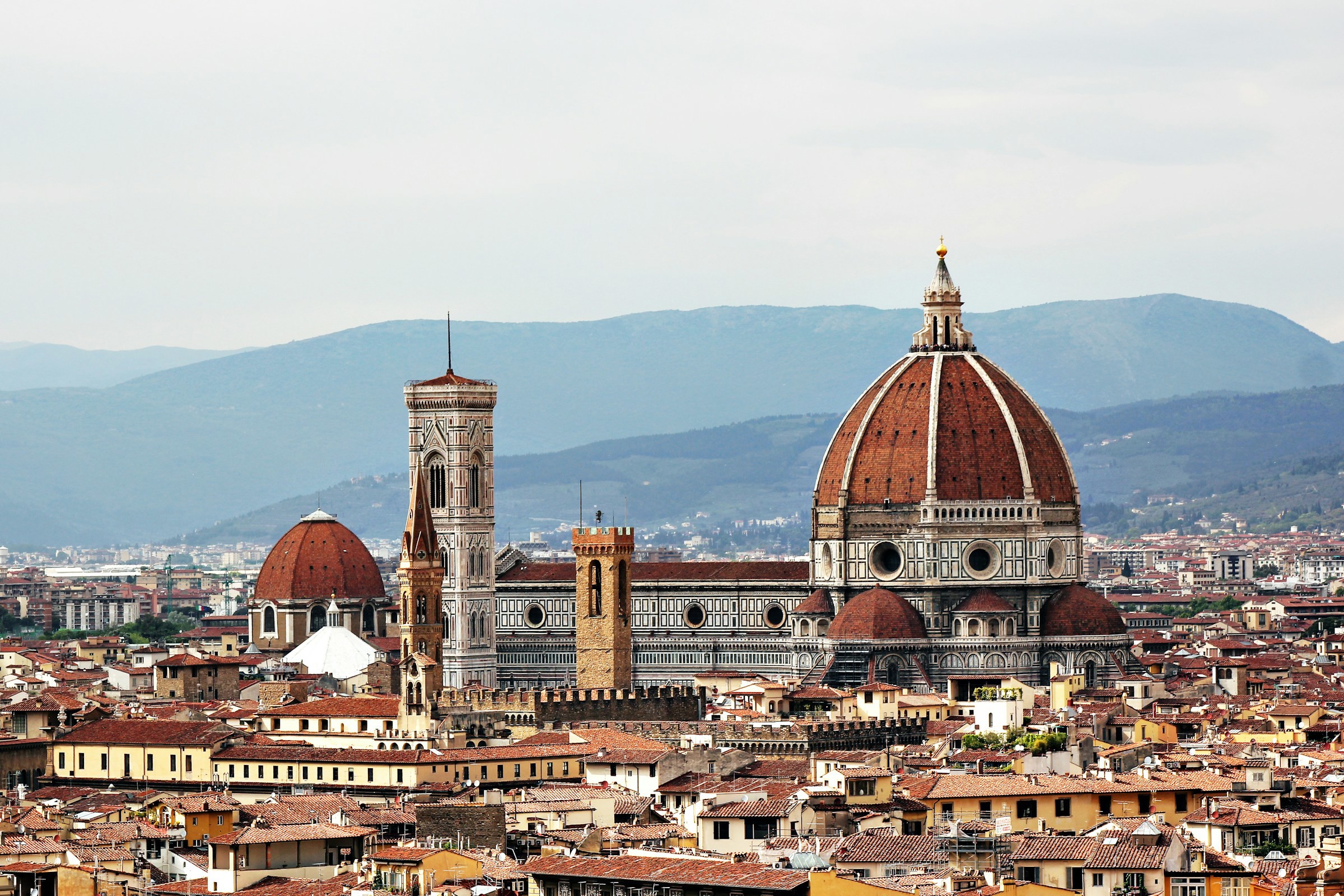 Utsikt over katedralen Duomo i Firenze, med byen rundt den og åser i bakgrunnen som møter en overskyet himmel