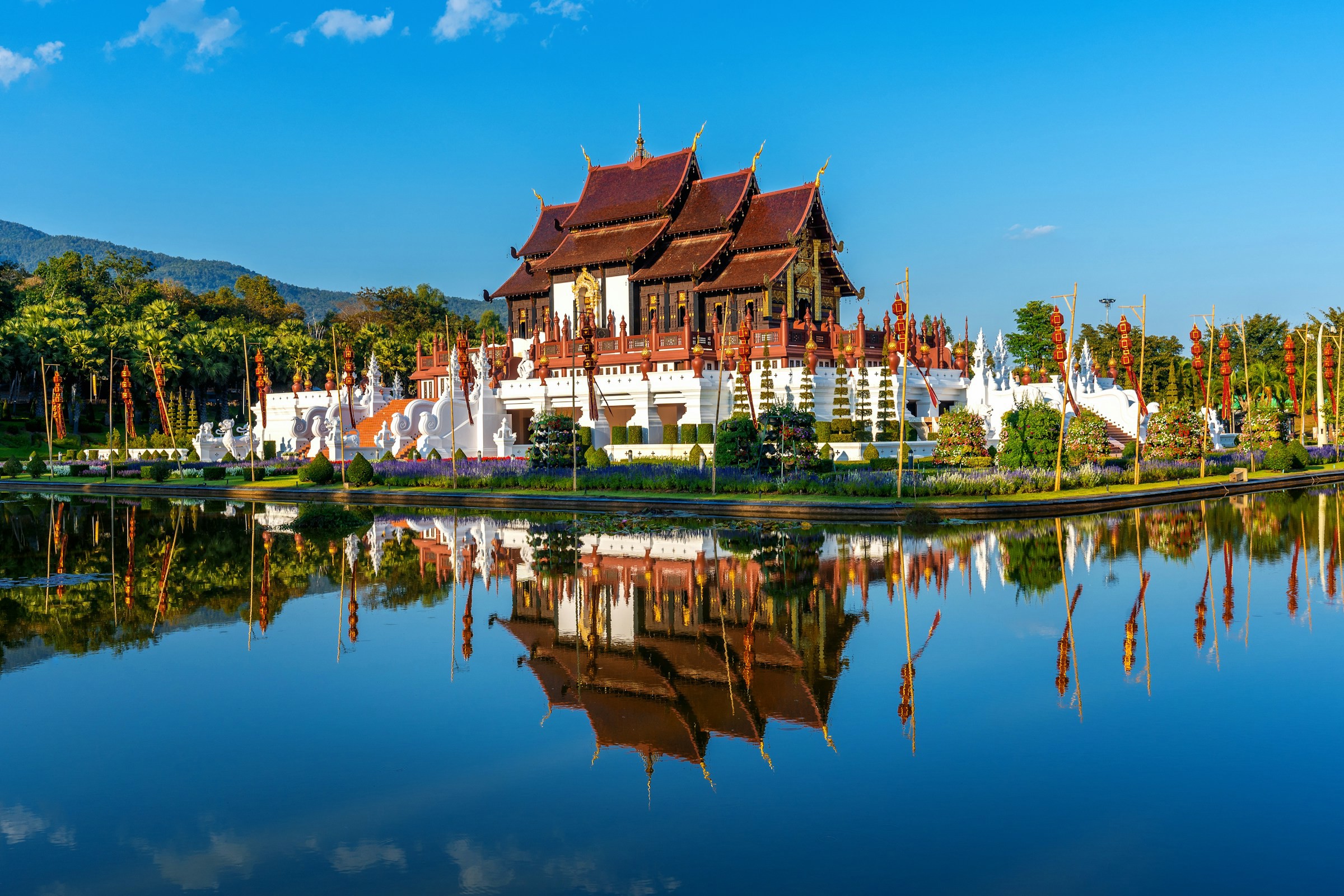 Templer i Phnom Penh med utsmykkede tredetaljer reflekteres i en vannoverflate, omgitt av frodige grøntområder og klar blå himmel.