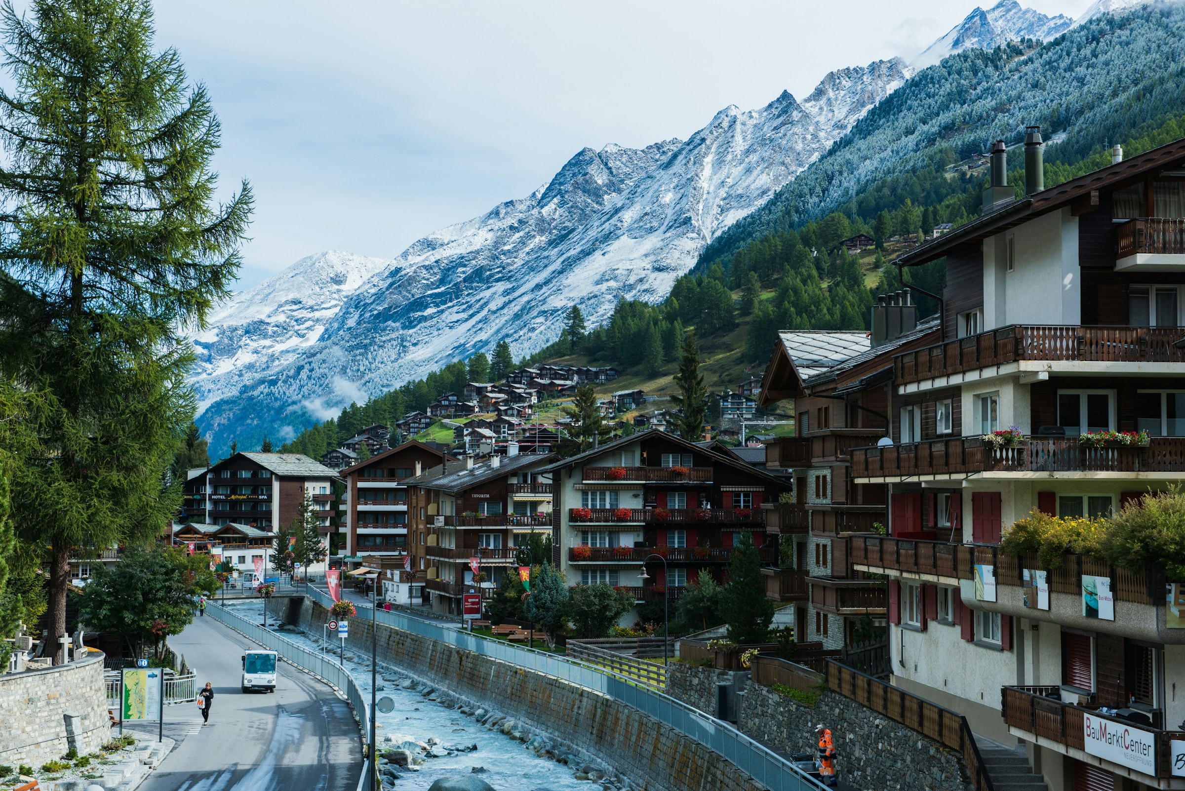 Swiss mountain scenery with traditional chalet-style buildings and a winding river surrounded by coniferous trees and snow-capped mountain peaks.