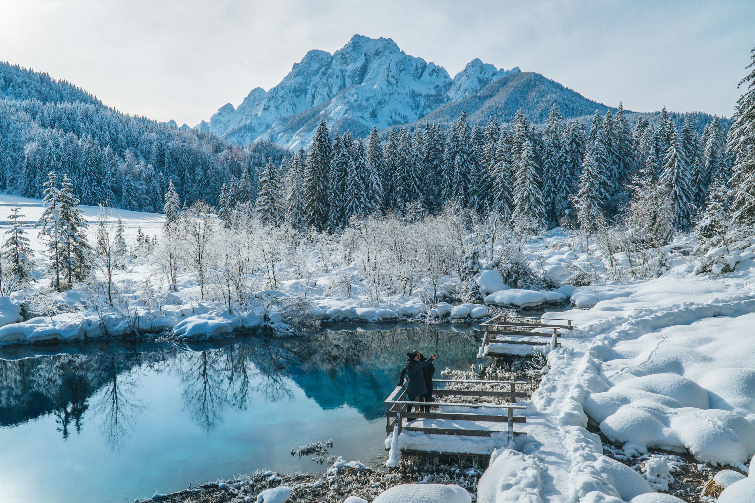 Two people standing by a lake in a snow-covered winter wonderland with forest and beautiful mountains in the background in Ranjska Gora