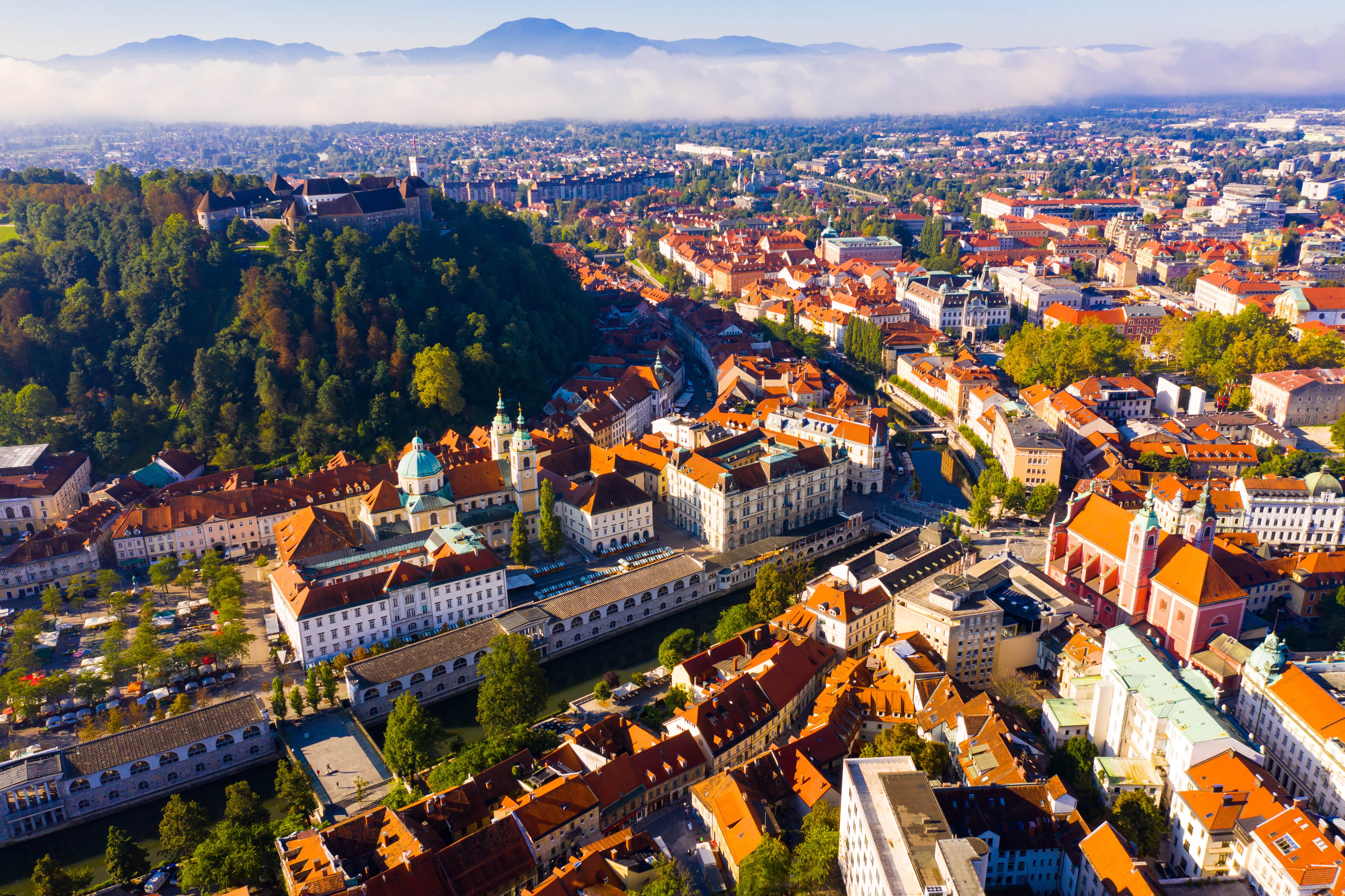 Travel to Ljubljana - Bird's eye view of city hall with lush forest next to it and beautiful mountains and clouds in the background