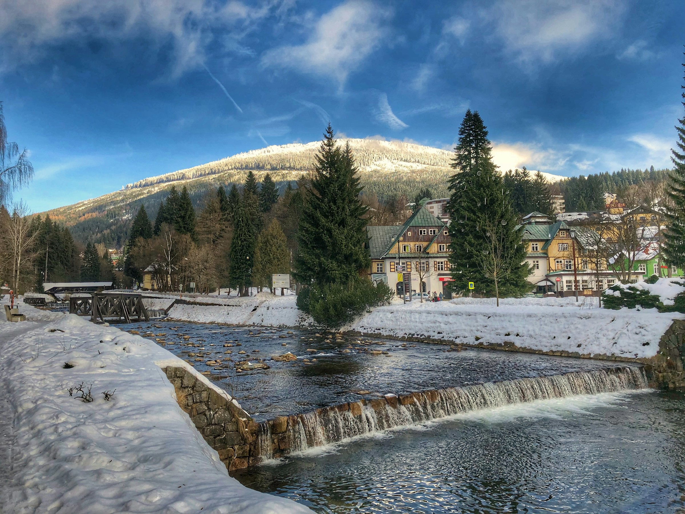 A river runs through a small snow-covered village in Spindleruv Mlyn with mountains and blue sky in the background