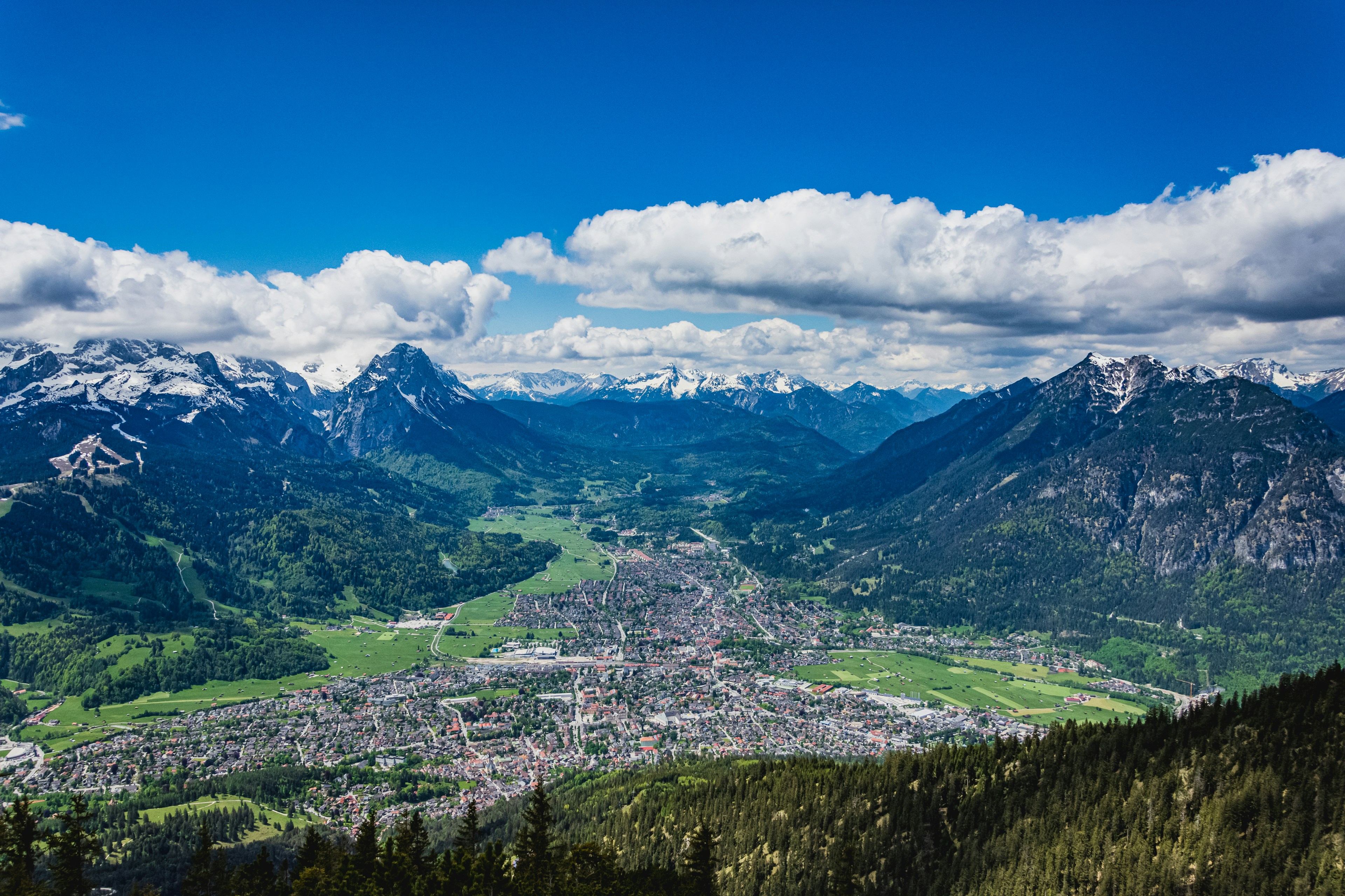 Travel to Garmisch - Panoramic view of the city between between forest and mighty snow-capped mountains with blue sky in the background