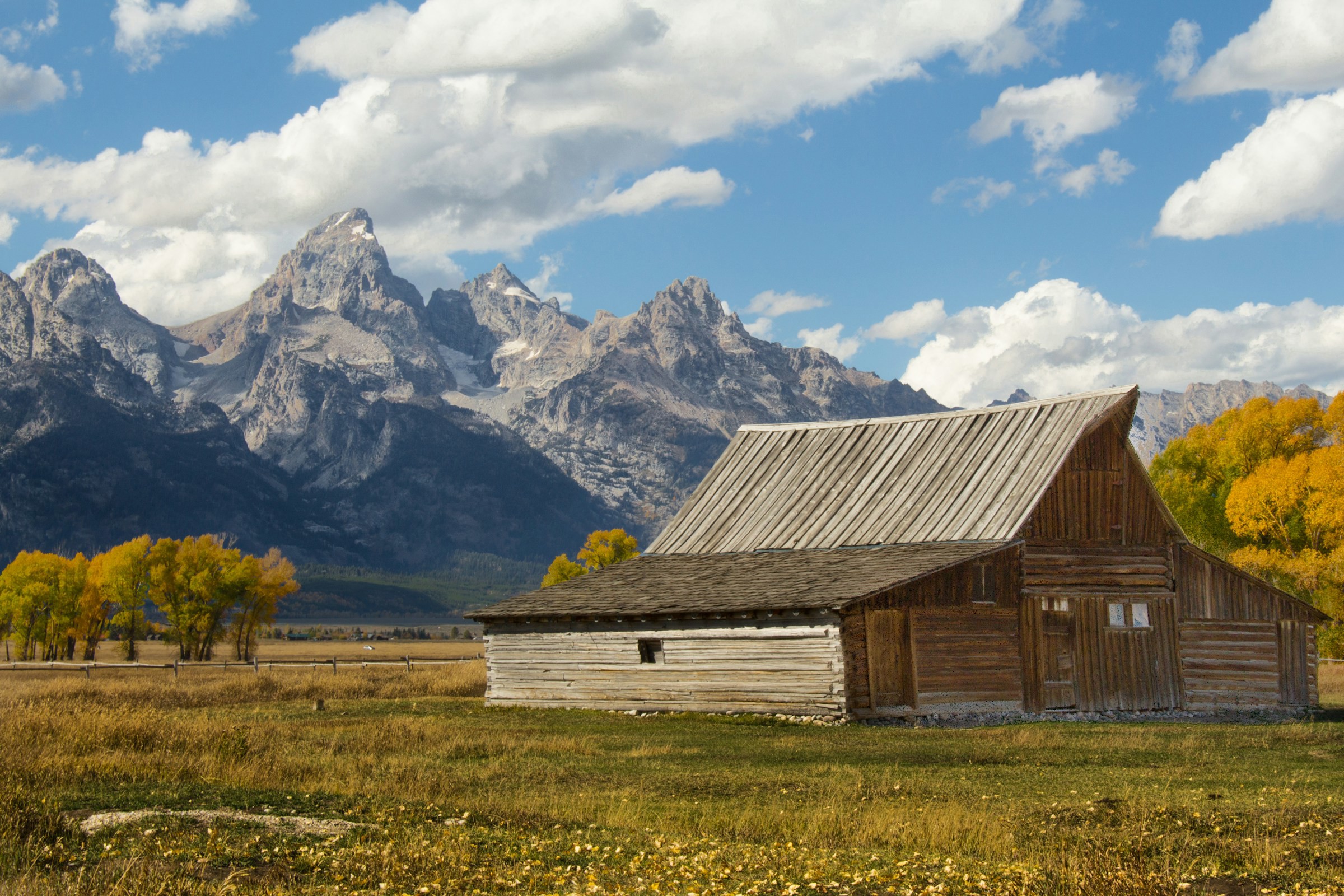 Old wooden barn in front of the Teton Mountains with autumn-colored deciduous trees and blue sky in the background.