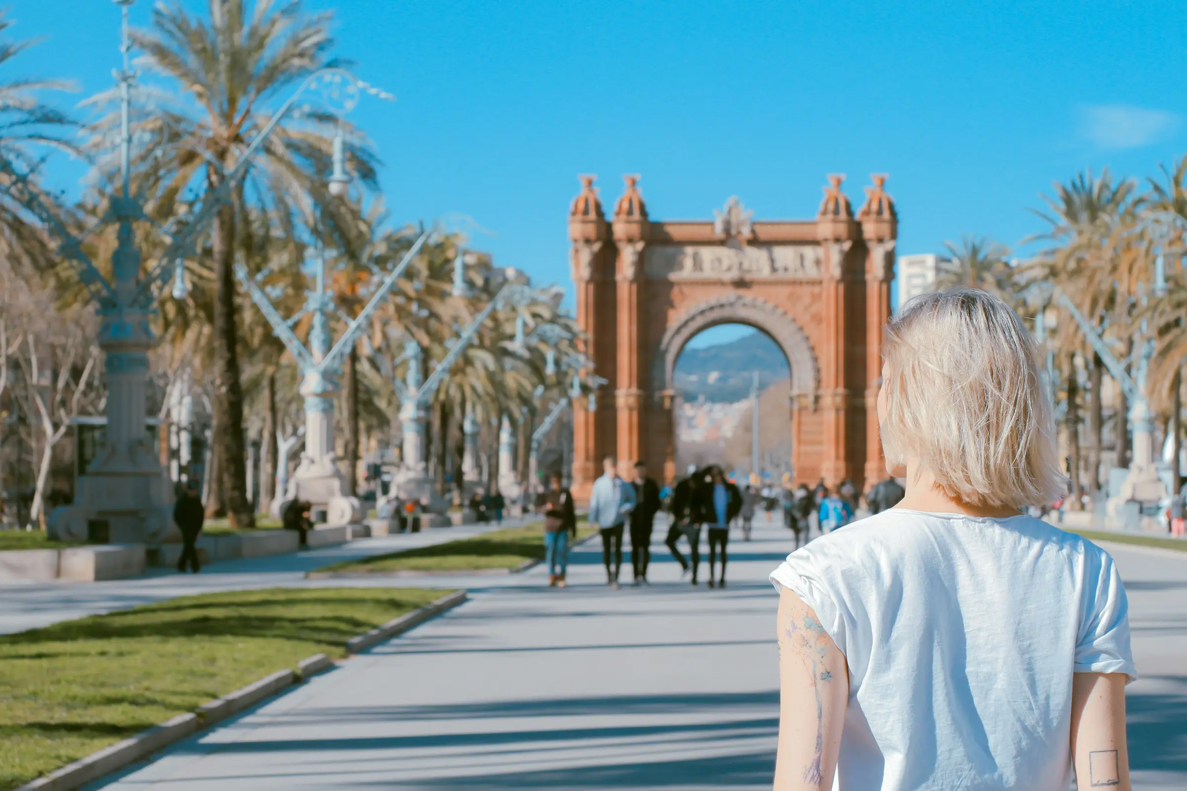 Blonde woman with tattoos facing the Arc de Triomf in Barcelona, with palm tree-lined promenade and pedestrians under a clear blue sky.