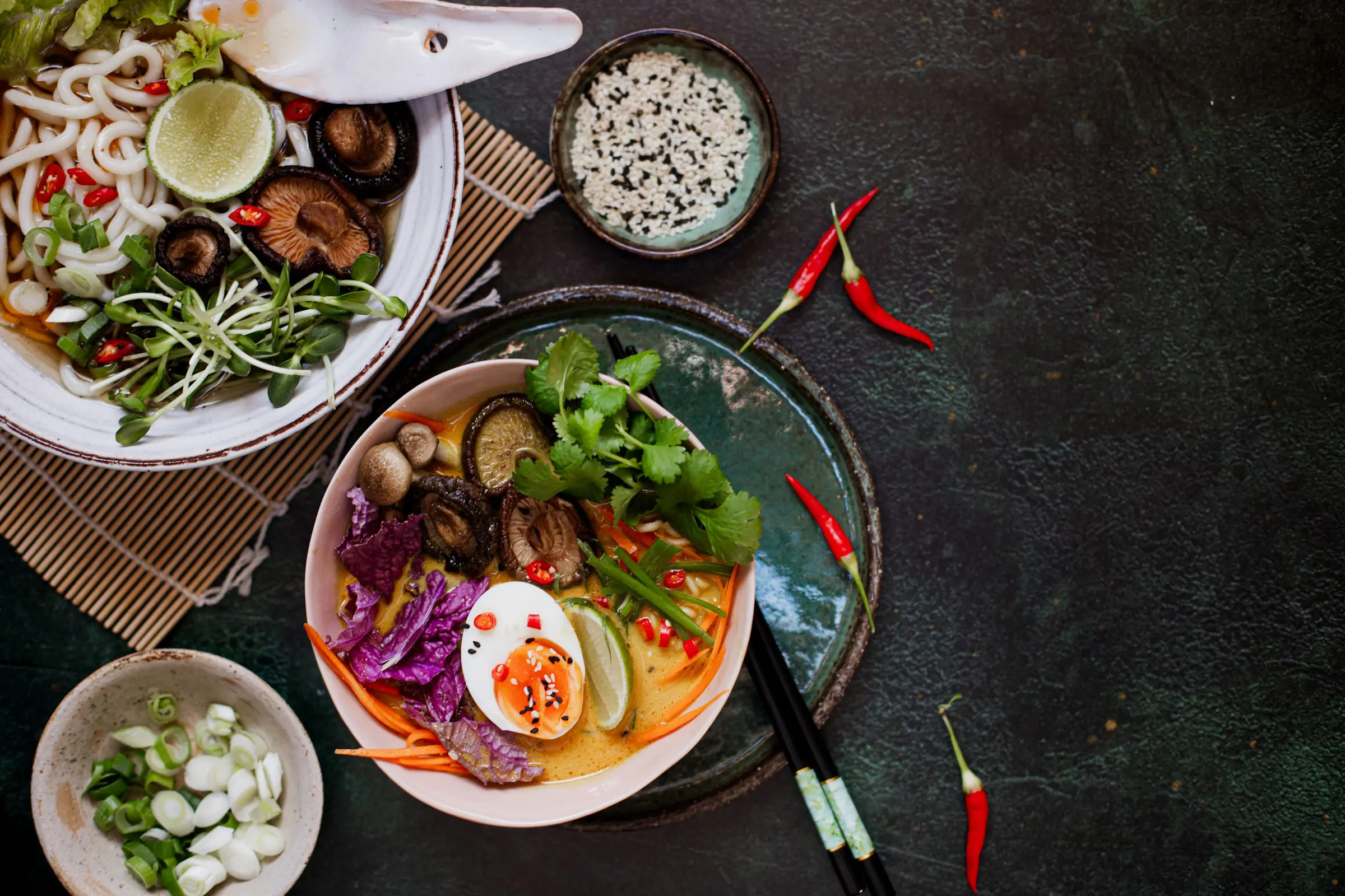 A vibrant display of Asian noodle soup featuring an assortment of fresh vegetables, boiled egg, mushrooms, lime wedges, and chili peppers, with garnishes of cilantro and green onions on the side.