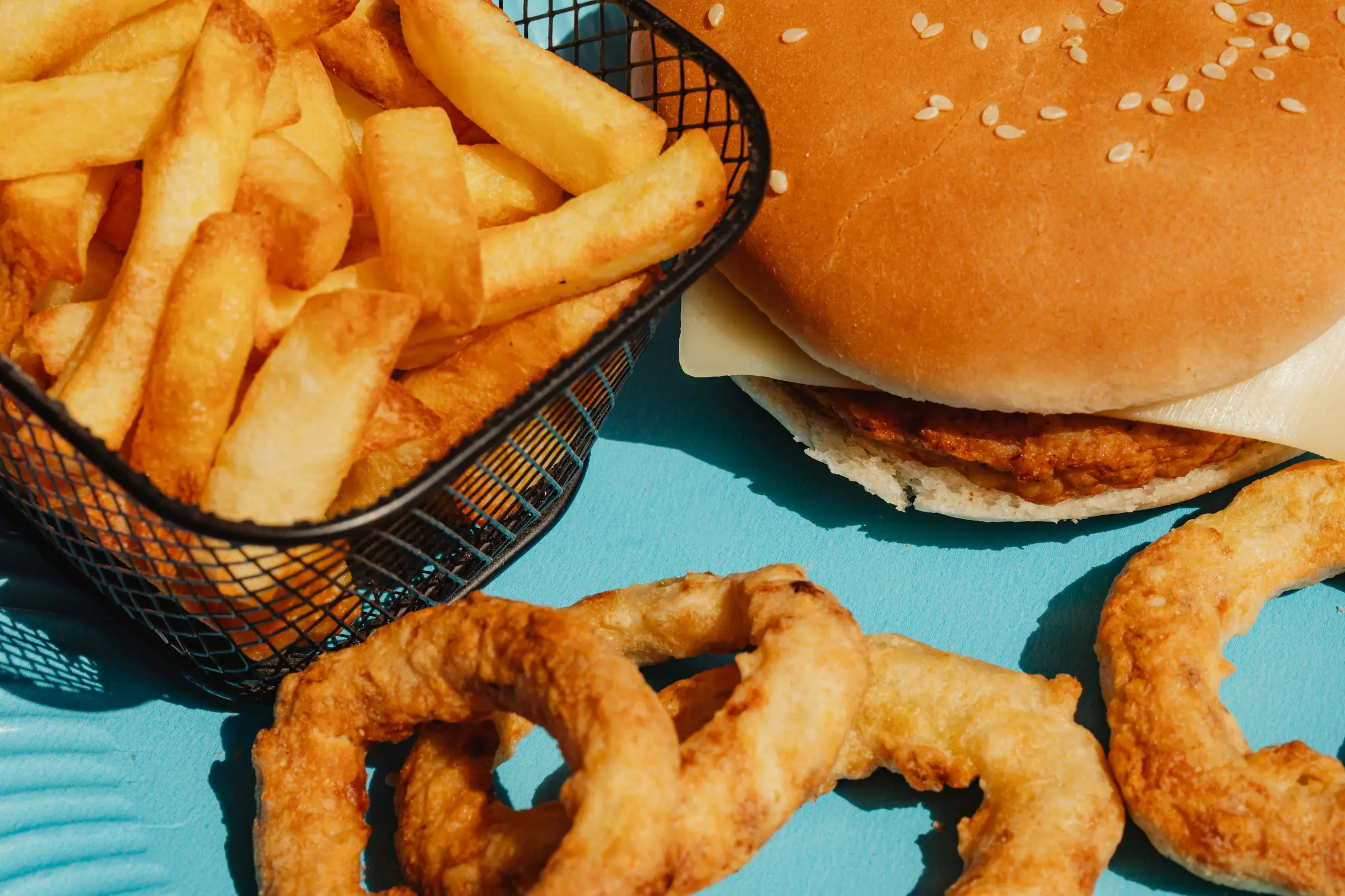 Burger with sesame seed bun, crispy French fries in a basket, and a side of golden-brown onion rings on a bright blue surface.