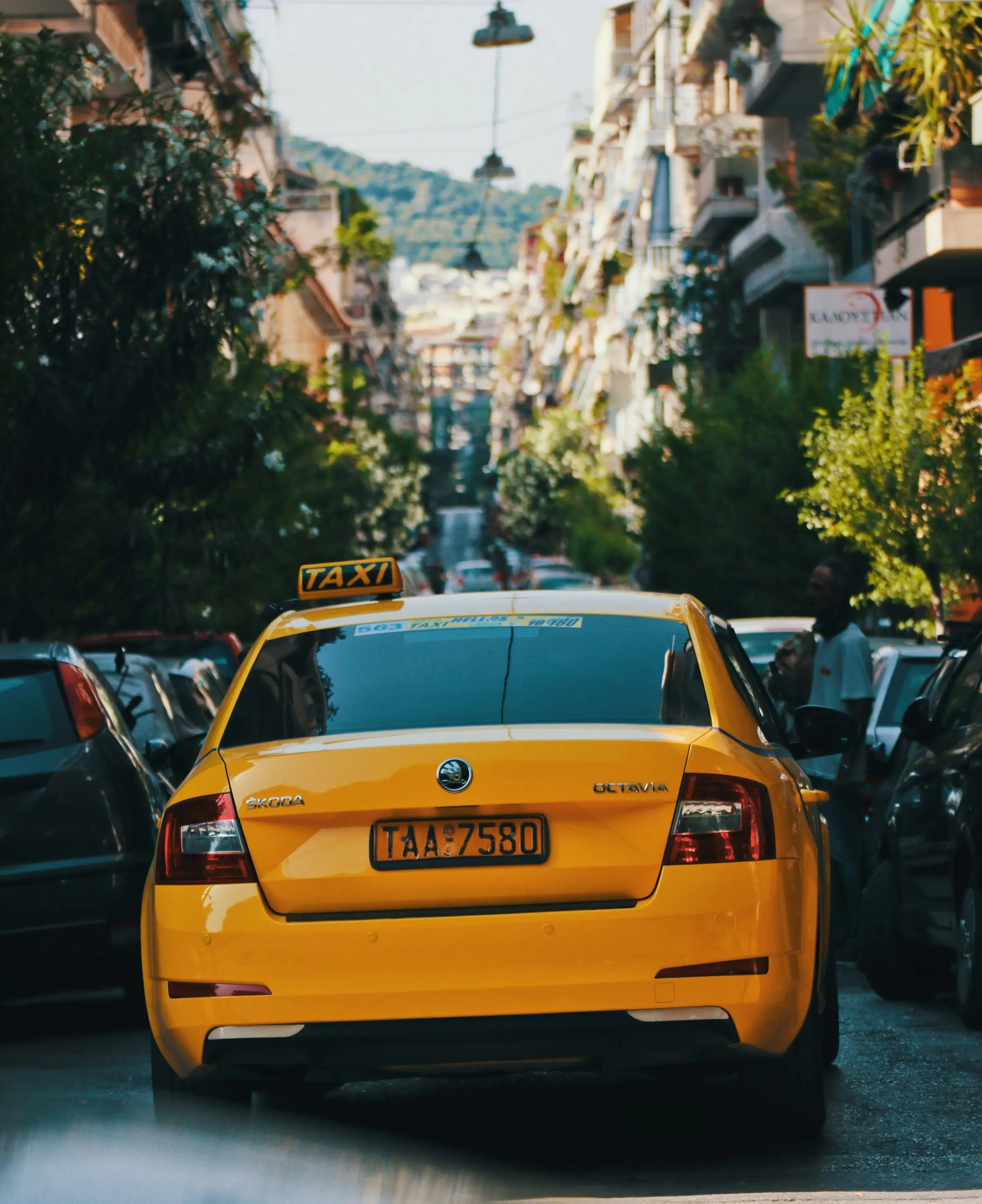 Bright yellow taxi driving down a narrow, bustling street lined with parked cars and apartment buildings, with a scenic hillside in the background.